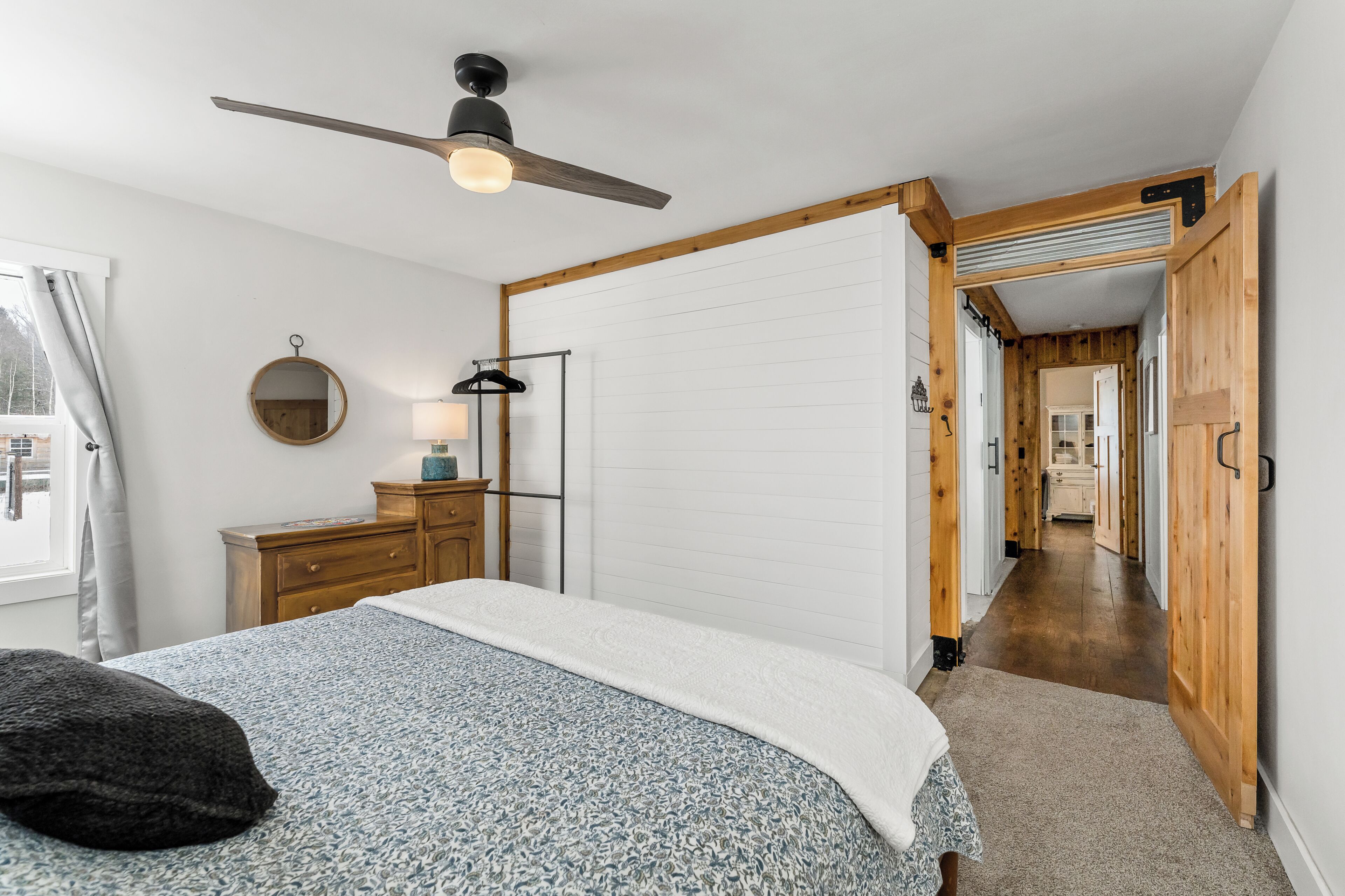 A bedroom with a bed, a ceiling fan, a dresser, a mirror, and a hallway leading to another room.