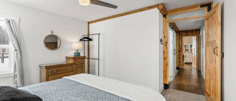 A bedroom with a bed, a ceiling fan, a dresser, a mirror, and a hallway leading to another room.