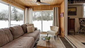 L’intérieur d’un chalet accueillant, avec un plafond en bois, un canapé, un fauteuil, une table basse agrémentée d’une plante, et une vue sur un paysage enneigé par de grandes fenêtres.