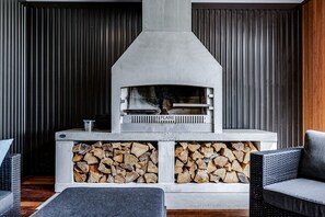 A fireplace with a stone facade, wood storage below, and wicker furniture. - Olive Grove • Relaxing Retreat • Rangitahi (Raglan)