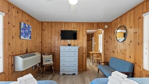 A room with wooden walls, a TV, a dresser, a chair, a sofa, and two beds with towels. - Snug Harbor Inn | Blueberry Suite | Near Lake MI (Muskegon)