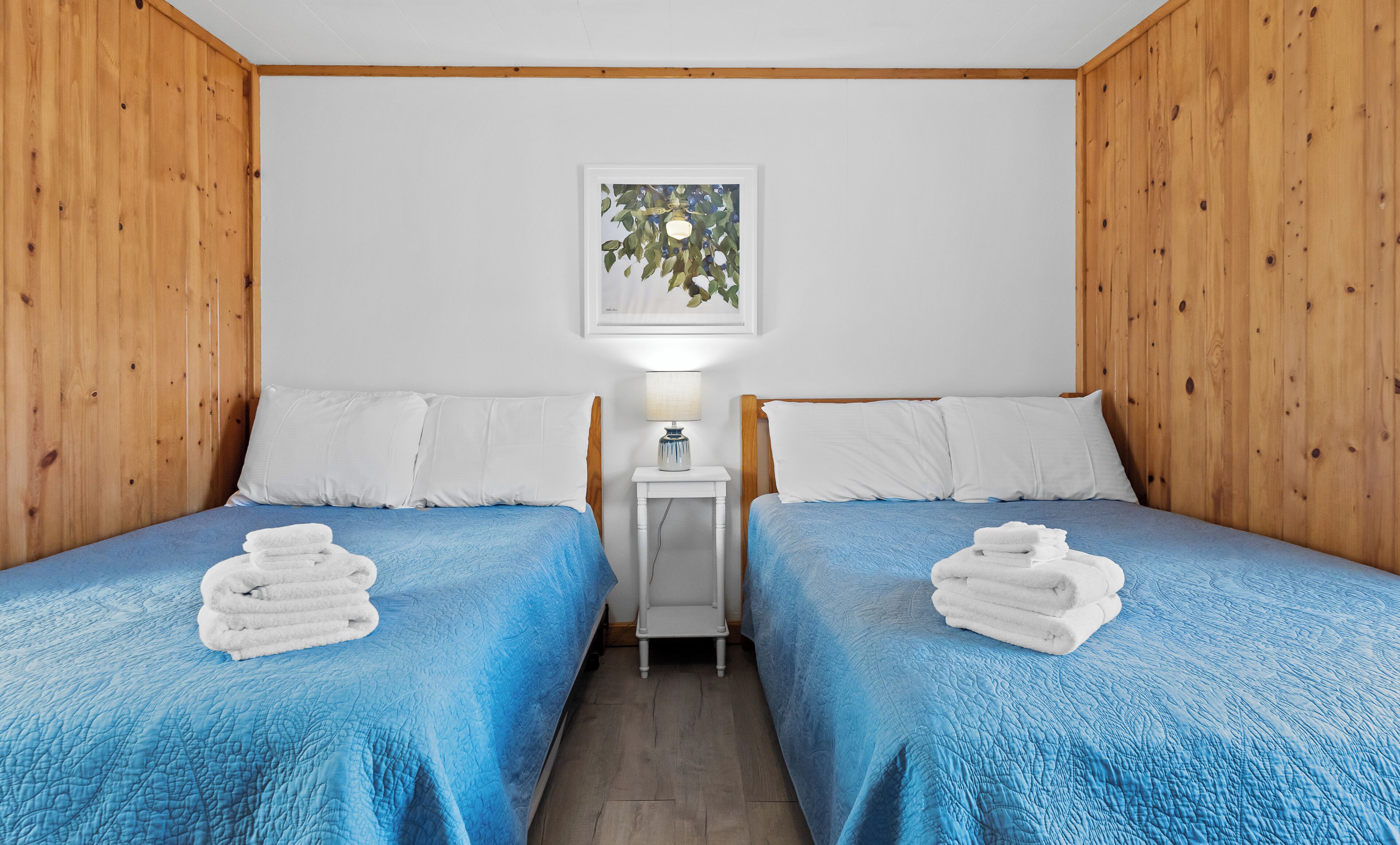 A room with two beds, white pillows, a small bedside table with a lamp, and a framed picture on the wall.