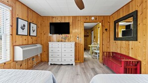 A room with wooden walls, a ceiling fan, a TV, a white dresser, a red bench, and two beds. - Snug Harbor Inn | The Roost | Near Lake MI (Muskegon)
