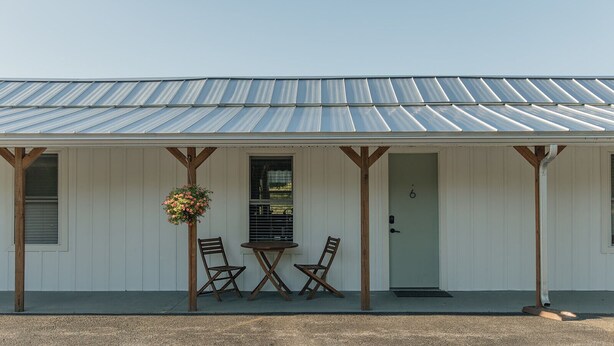 A white building with a metal roof, a porch with two wooden chairs, and a hanging flower basket. - Snug Harbor Inn | The Roost | Near Lake MI (Muskegon)