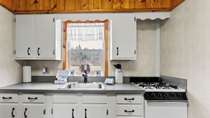 A kitchen with white cabinets, a gas stove, a sink, and a window with a view of trees. - Snug Harbor Inn | The Roost | Near Lake MI (Muskegon)