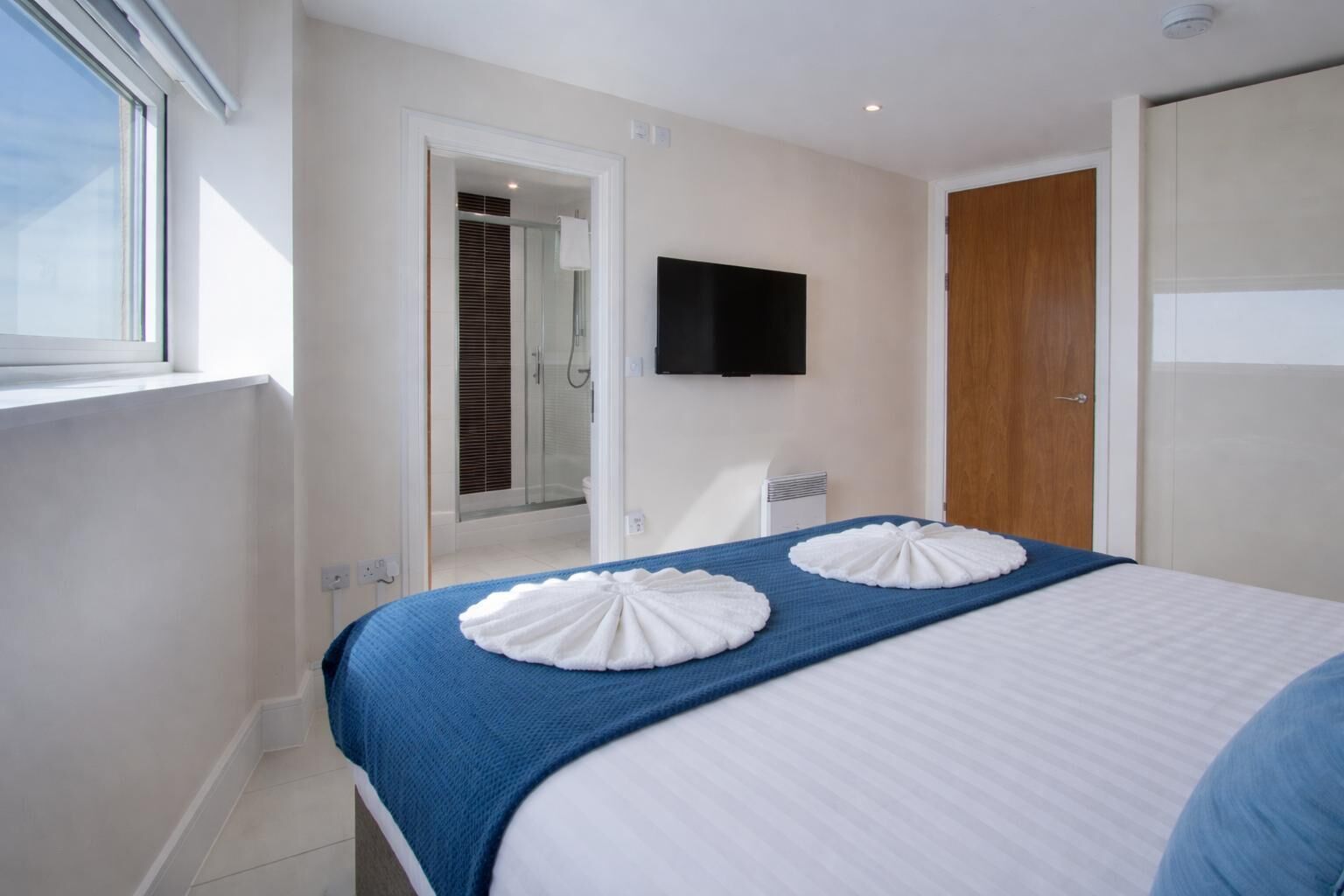 A hotel room with a bed, a TV mounted on the wall, a shower area, and a wooden door.