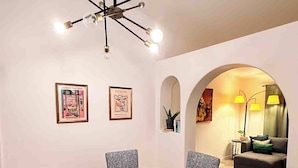 Un coin repas avec une table ronde en bois et six chaises, deux œuvres d’art encadrées accrochées au mur, et un espace de vie accueillant visible à travers une arche.