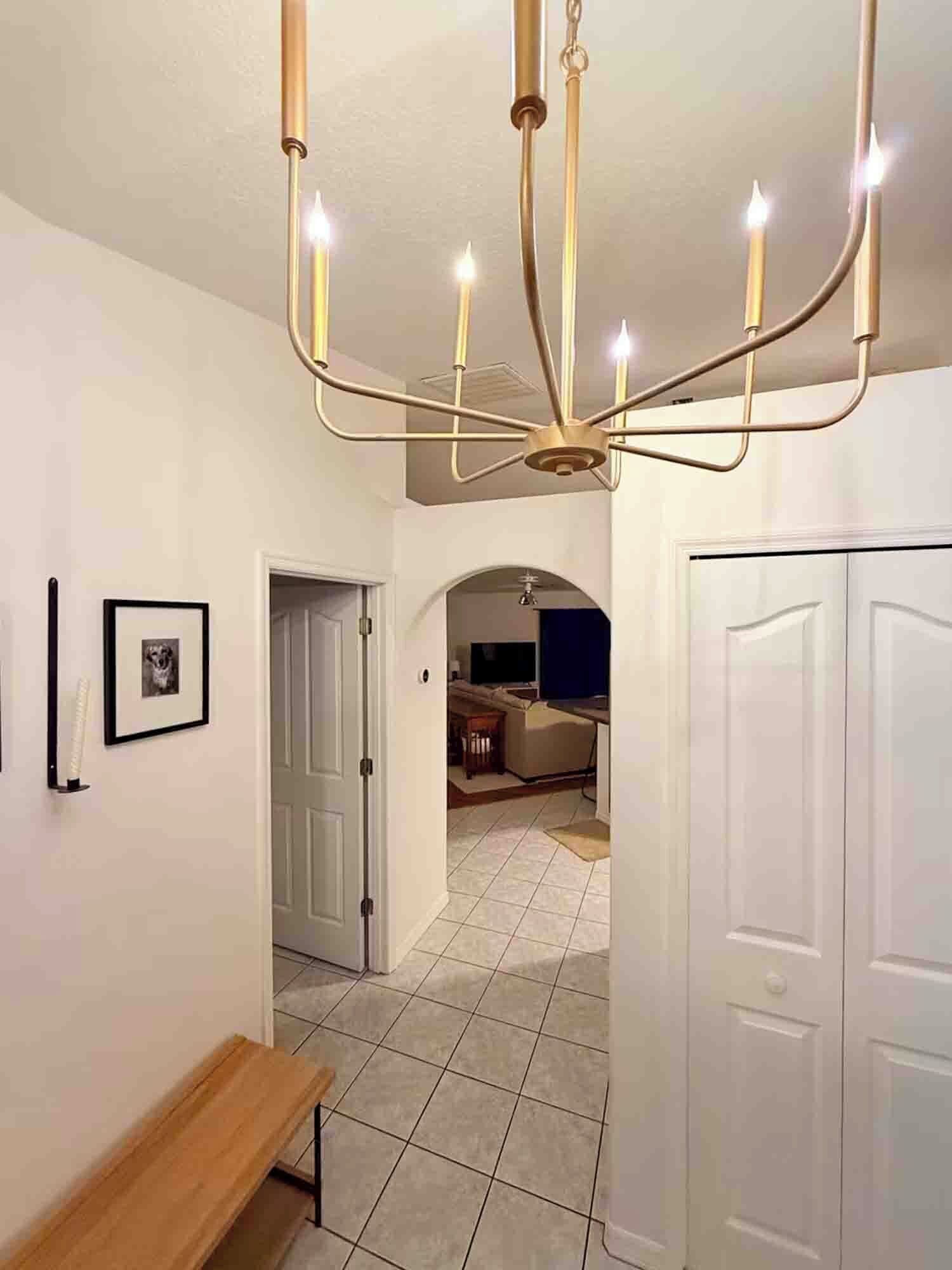 A hallway with a chandelier, a bench, a framed picture, and a door leading to a room.