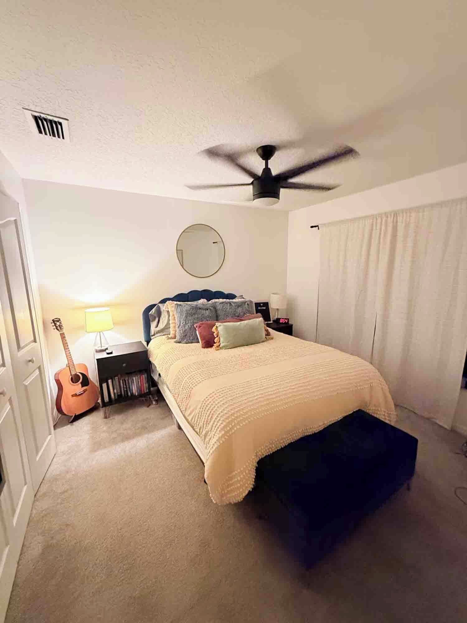 A bedroom with a bed, a ceiling fan, a guitar, a bedside table, and a lamp.