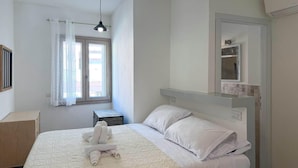 Ein Schlafzimmer mit einem Bett, einem Nachttisch, einem Fenster mit Vorhängen, einer Hängelampe und einem fest installierten Regal über dem Bett.