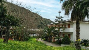 A building with a white exterior, surrounded by greenery and a hillside. - 2 bedroom lovely apartment in Ricadi (Ricadi)