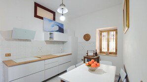 Una cucina con mobili bianchi, un piano di lavoro in legno, un tavolo bianco con una ciotola di arance, una finestra con tende decorative e una lampada a soffitto.