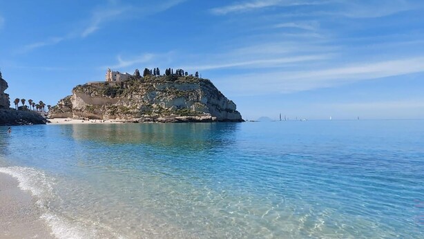 Paesaggio costiero con acque limpide, una spiaggia sabbiosa e un promontorio roccioso con un edificio sulla cima.