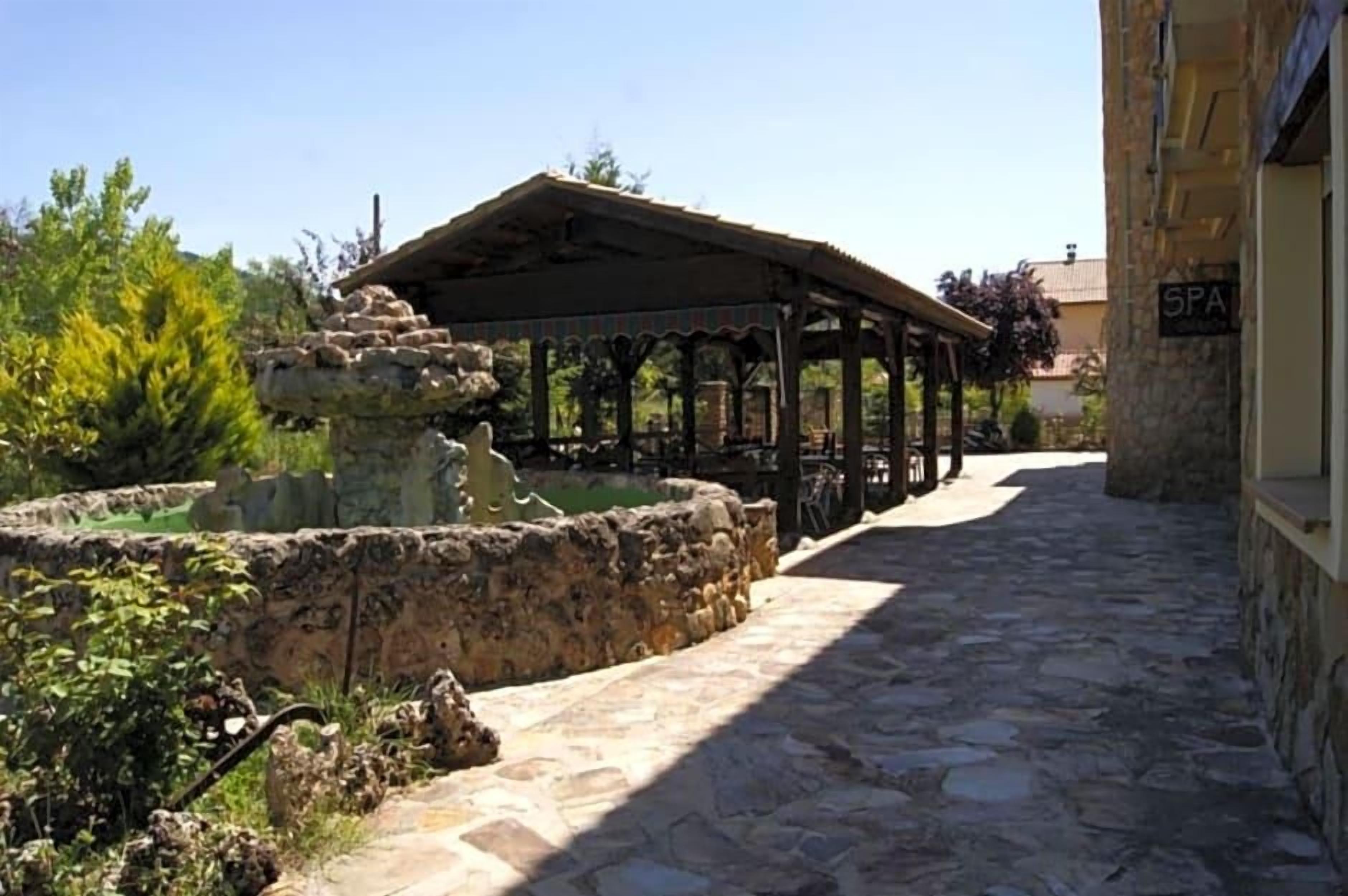 Un camino de piedra conduce a un espacio al aire libre cubierto, con una fuente, un letrero de spa y vegetación.