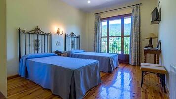 A room with two beds, a window with curtains, a radiator, and a small table with a lamp.