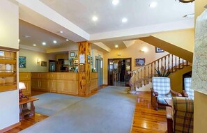 Un hall d’entrée spacieux doté d’un plancher en bois, d’un comptoir de réception, d’un escalier et d’un espace salon avec des fauteuils à carreaux.