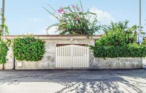 A gated entrance with a concrete wall, a metal gate, and green foliage with pink flowers. - Cozy home in S. Maria del Focallo with WiFi (S. Maria del Focallo)