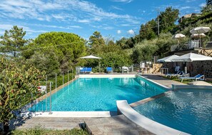 A rectangular swimming pool with a stone deck, surrounded by lounge chairs and umbrellas, set against a backdrop of trees and a stone building. - Awesome apartment in Montecastelli Pisano (Montecastelli Pisano)
