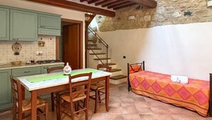 Una stanza accogliente con un tavolo da pranzo, sedie, un letto con coperta colorata, angolo cottura con pensili, fornello e una scala che conduce a un piano superiore.