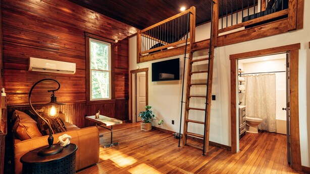 A spacious room with wooden floors, a loft area with a wooden staircase, and a bathroom with a shower curtain. - Corton Lodge – Riverfront Wedding & Group Retreat (Clendenin)