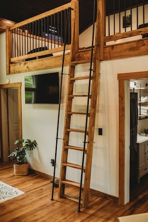 A loft-style room with a wooden ladder leading to a balcony, a flat-screen TV mounted on the wall, and a potted plant on the floor.