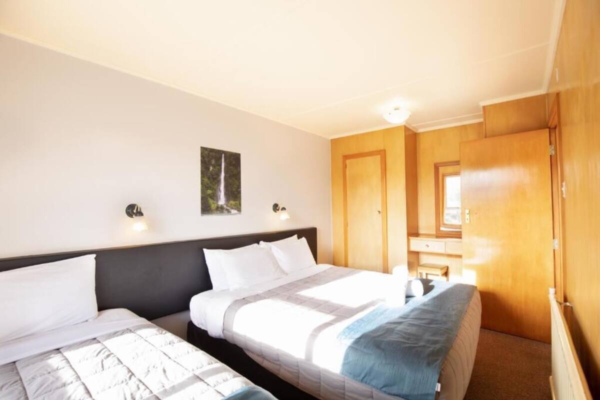 Habitación de hotel con dos camas, un armario de madera, un escritorio con silla y una lámpara fijada a la pared.