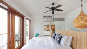 Une chambre à coucher avec un lit, un ventilateur de plafond, un luminaire suspendu et une vue sur l’extérieur, accessible par une porte coulissante.