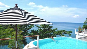 Une piscine à débordement offrant une vue sur l’océan, un parasol à rayures et une rambarde blanche.