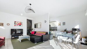A modern living room with a flat-screen TV, a sofa, a coffee table, and a dining area with a glass table and chairs.