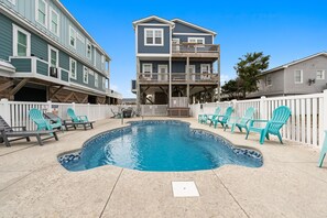A swimming pool surrounded by turquoise chairs, a white picket fence, and a multi-story building with balconies. - Done Dune Stuff - Experience beachside bliss at a 4-bedroom retreat in Oak Island! (Oak Island)