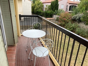 A balcony with a round table and two chairs, a tiled floor, and a view of a garden with various plants and a building in the background. - 2-room apartment, terrace - 4 guests (Argelès-sur-Mer)
