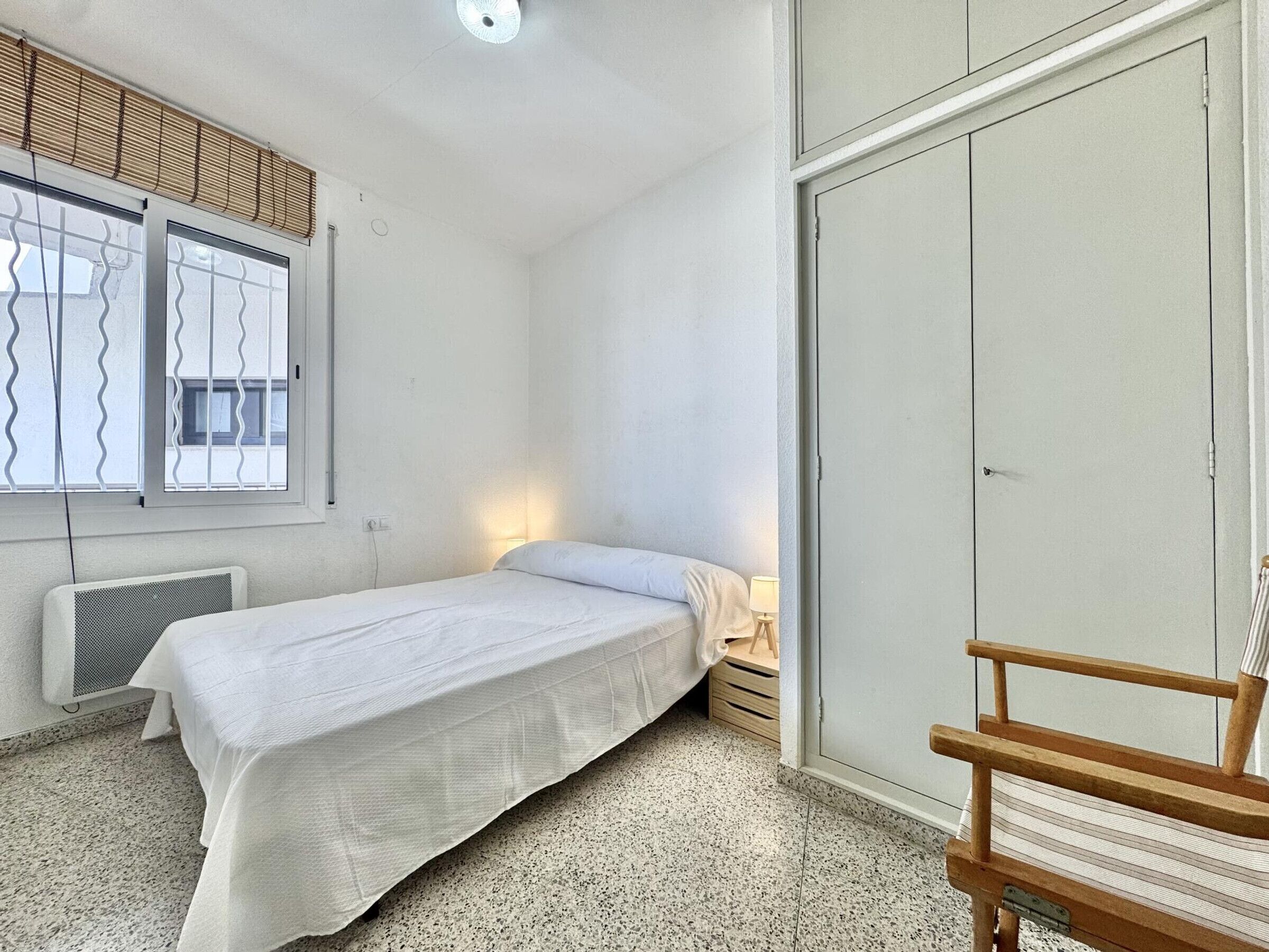 A bedroom with a bed, a chair, a wardrobe, a window with blinds, and a radiator.