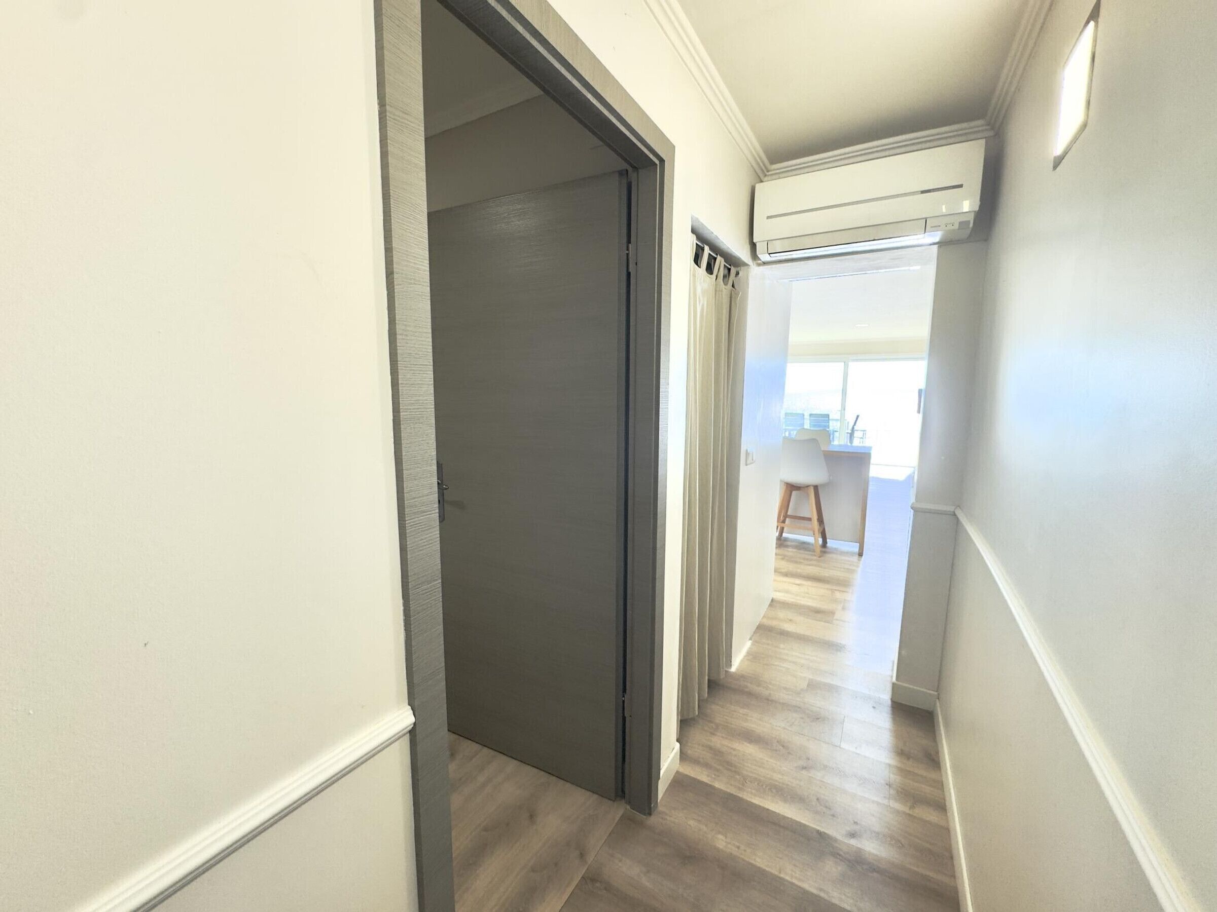 A hallway with a door, a ceiling-mounted air conditioner, and a view into a dining area with a table and chairs.