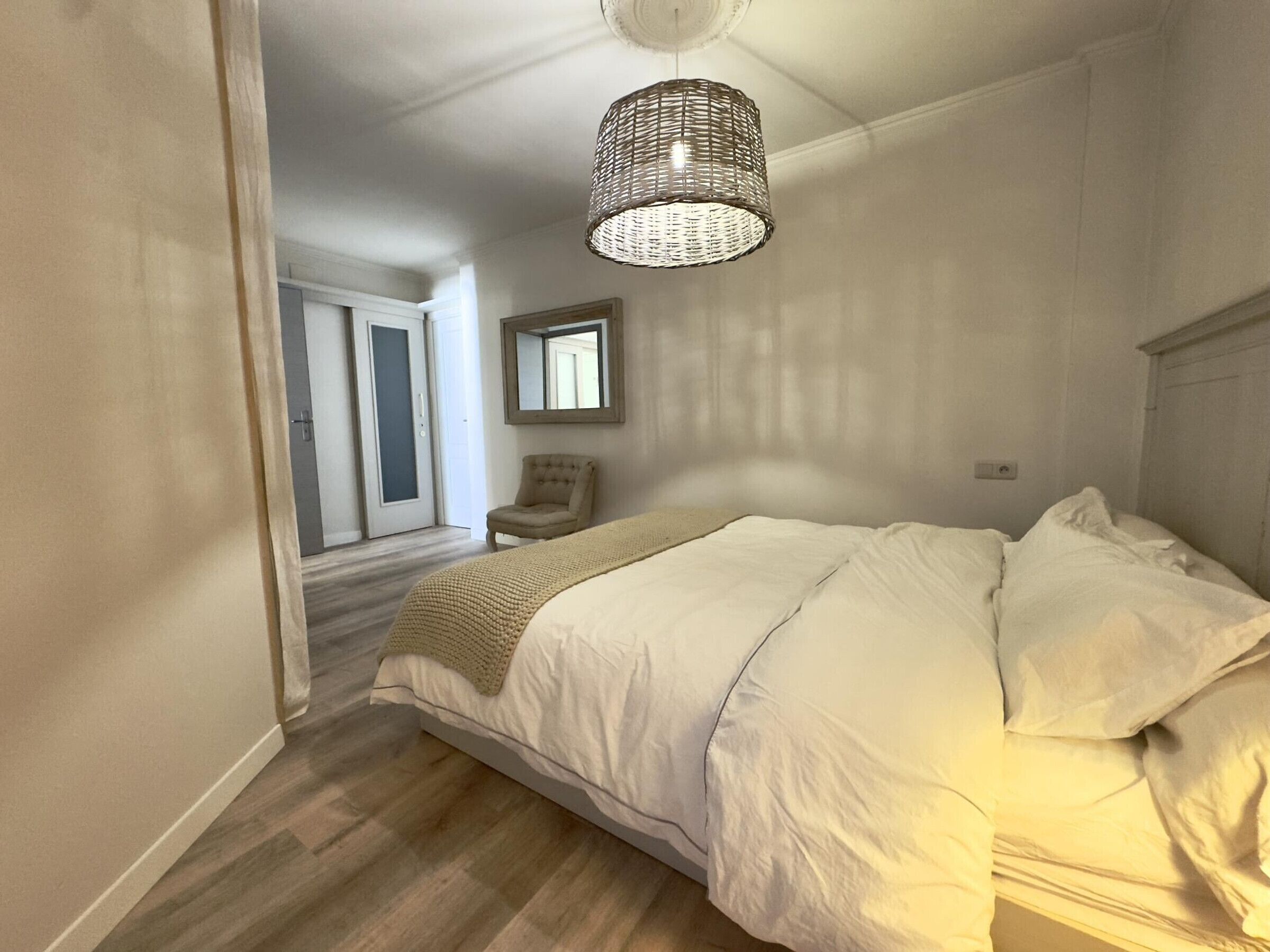 A bedroom with a large bed, a hanging light fixture, a chair, and a door leading to another room.