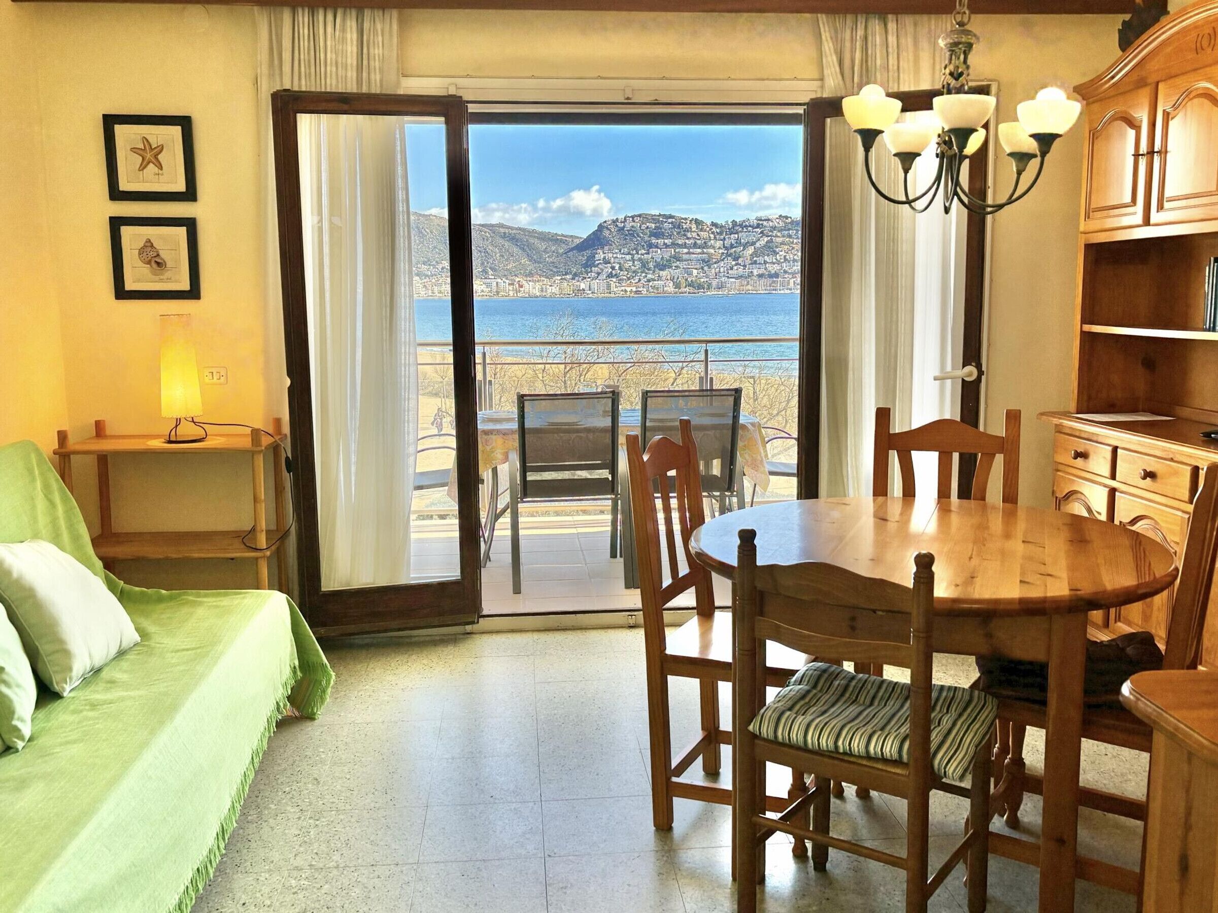 A room with a wooden dining table, chairs, a green sofa, a lamp, and a view of the sea through a large window.