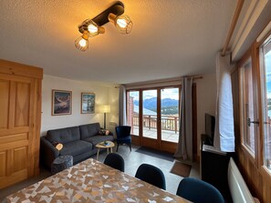 Quarto com sofá, mesa de jantar, televisão e varanda com vista para as montanhas.