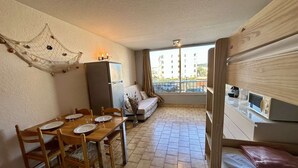 A compact room with a dining table, chairs, a sofa, a small kitchen area with a microwave, and a balcony with a view of buildings. - Practical studio cabin near beach - 4 guests (Cap d'Agde)