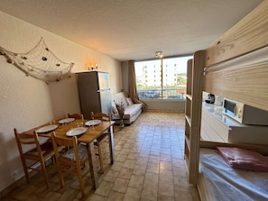 A compact room with a dining table, chairs, a sofa, a small kitchen area with a microwave, and a balcony with a view of buildings. - Practical studio cabin near beach - 4 guests (Cap d'Agde)