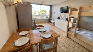 A compact room with a dining table set for four, a bunk bed, a small kitchenette, and a TV mounted on the wall. - Practical studio cabin near beach - 4 guests (Cap d'Agde)