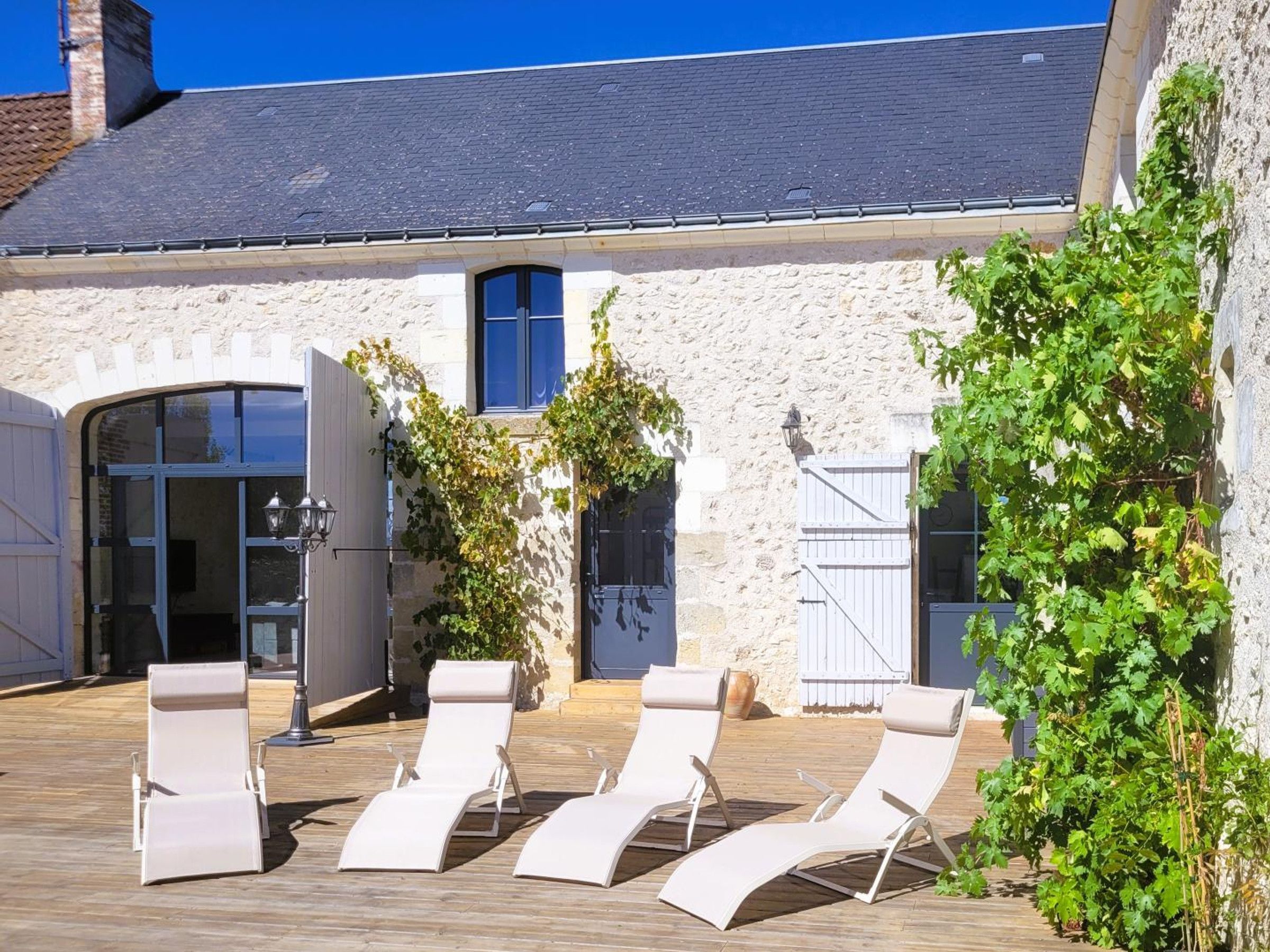 A courtyard with a stone building, a wooden deck, and three white lounge chairs.