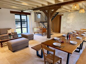 A spacious living area with a wooden dining table set for six, a sofa, a coffee table, and a staircase leading to an upper level. - Cottage with air conditioning - 9 guests (Le Louroux)