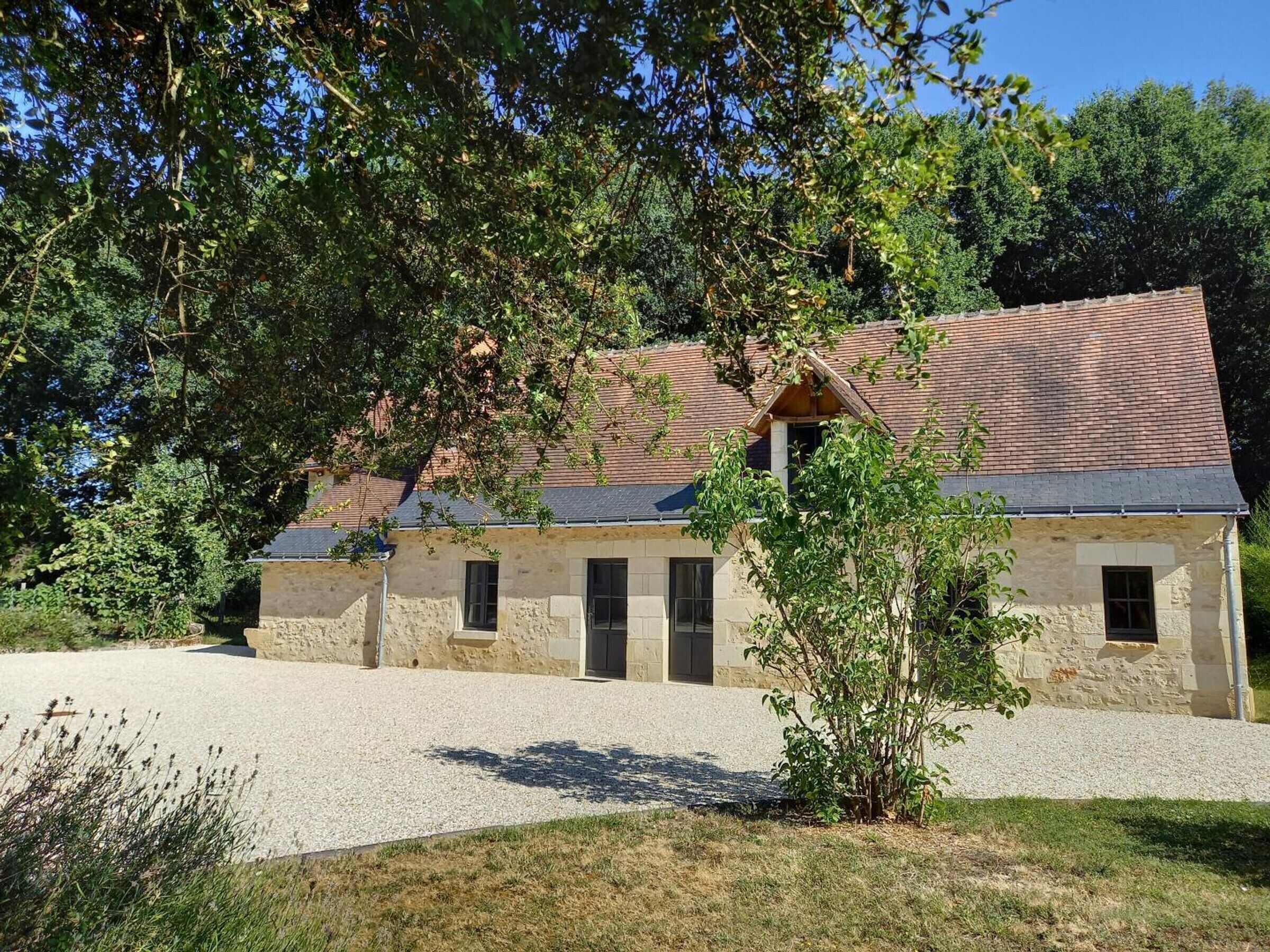 A stone building with a tiled roof, a gravel driveway, and surrounding greenery.