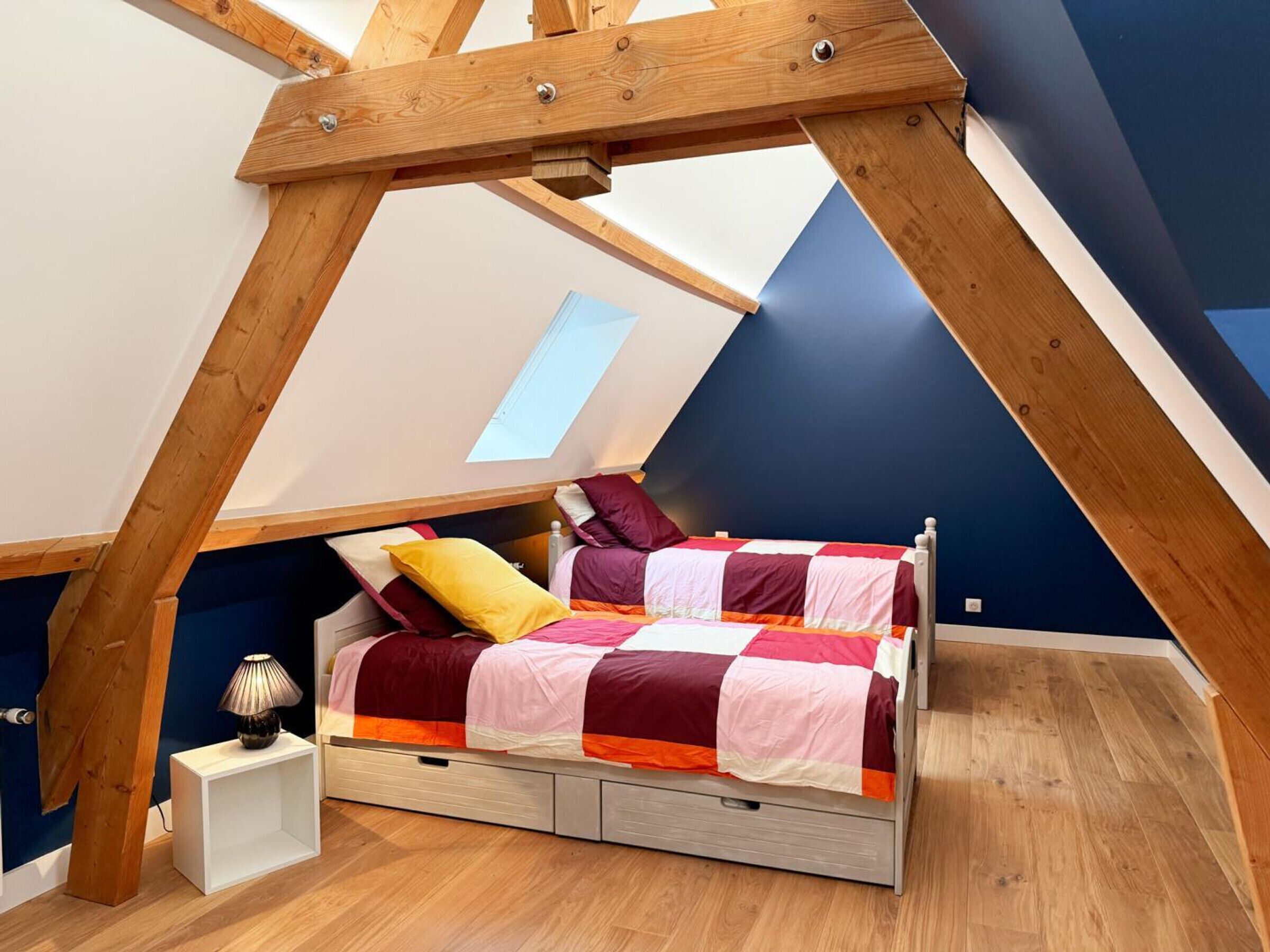 Ein Schlafzimmer im Loftsstil mit schrägem Dach, einem Oberlicht, einem Bett mit farbenfroher Decke und einer Nachttischlampe.