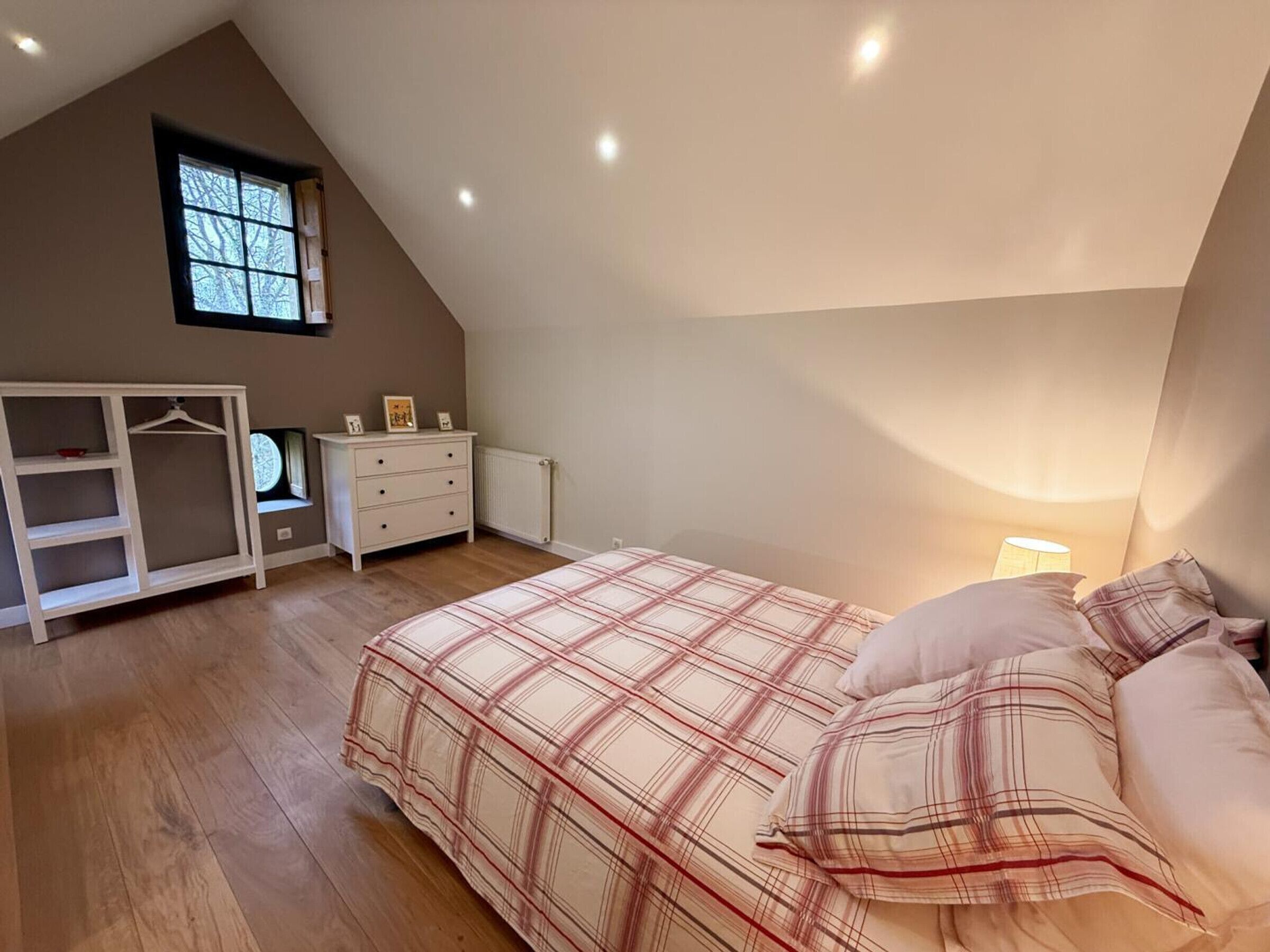 A bedroom with a sloped ceiling, a bed with plaid bedding, a dresser, a wardrobe, and a window.