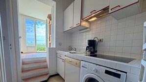 Cocina compacta con muebles blancos, lavadora, cafetera y vista al exterior por una puerta.