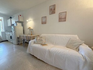 Ein helles Wohnzimmer mit einer Couch, einem Tisch und einer Waschmaschine. An der Wand befinden sich zwei gerahmte Bilder.