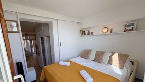 Un dormitorio con una cama, una mesita de noche, una estantería con libros y una puerta que da a otra habitación.