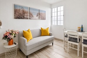 A modern living room with a grey sofa, yellow cushions, a dining table with chairs, and a window with blinds.