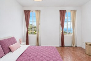 A bedroom with a bed, two windows with curtains, a nightstand, and a view of the sea.