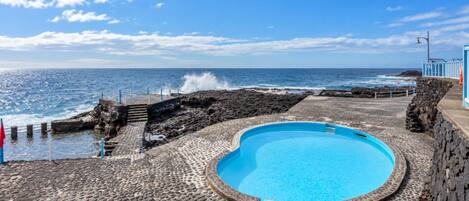 A pool with a stone pathway leading to the sea, a stone wall, and a clear view of the ocean.